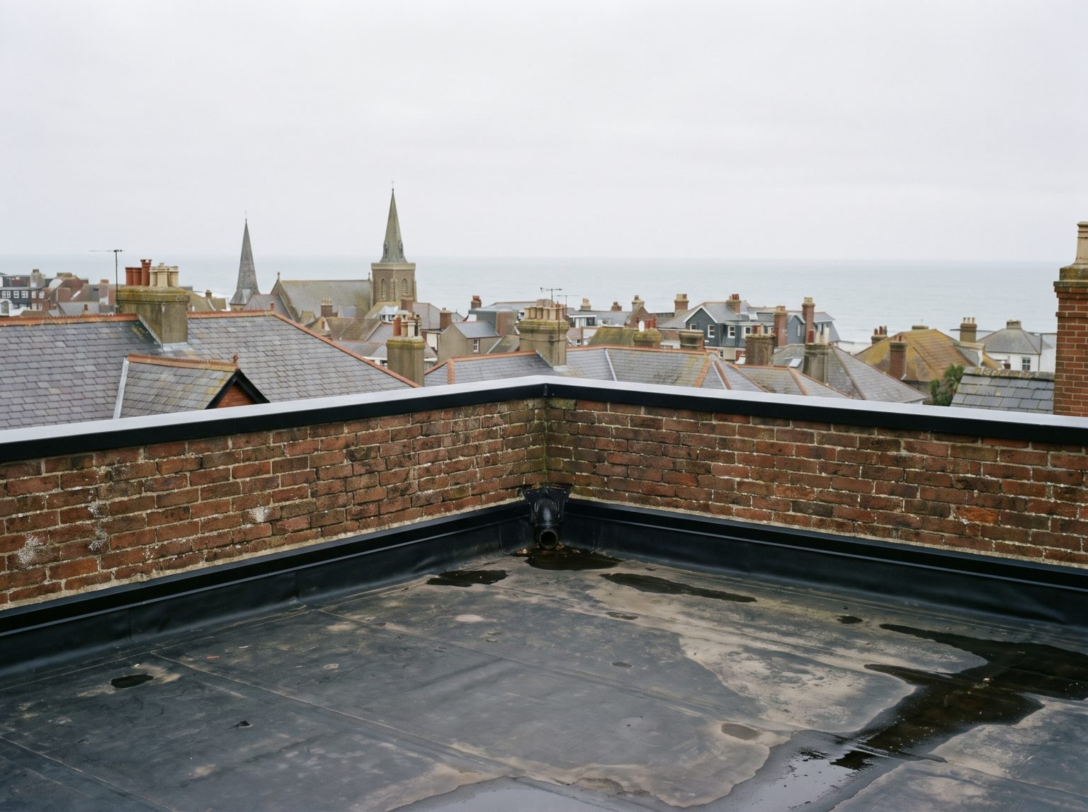A flat roof behind a low brick parapet on the top storey of a tall building, rooftops and sea beyond